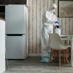 Person in hazmat suit disinfecting a modern kitchen table with spray bottle for hygiene.